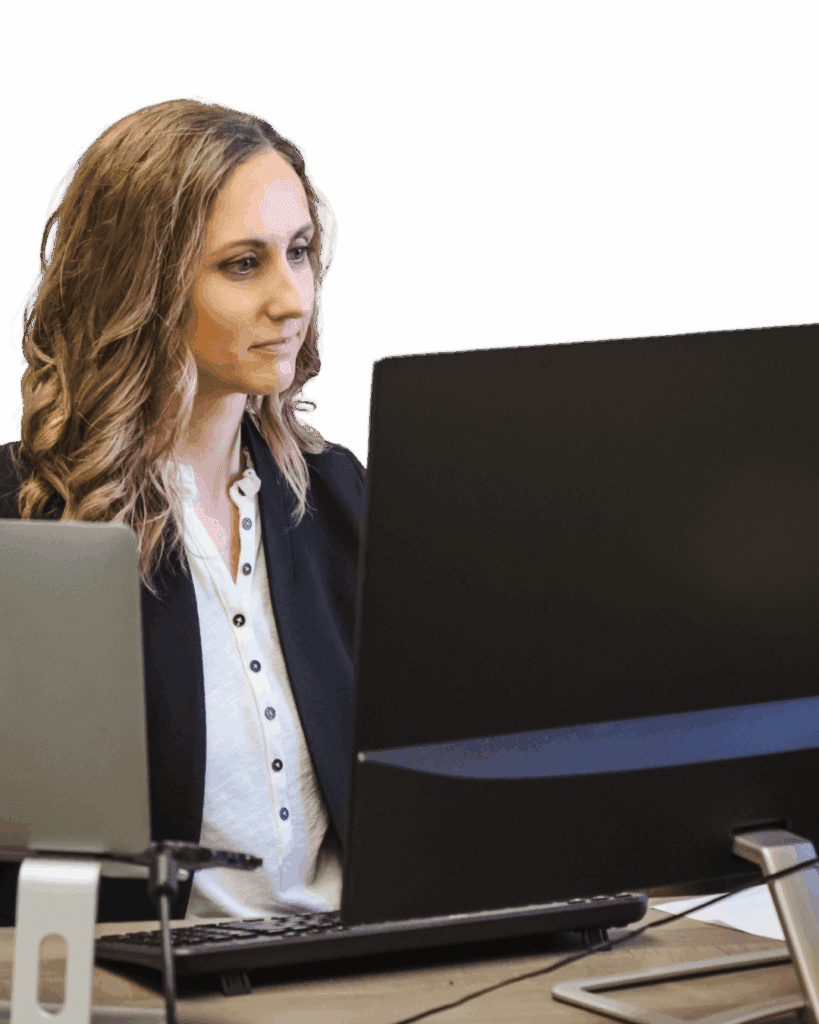 Woman with long wavy hair working at dual monitors at a desk.